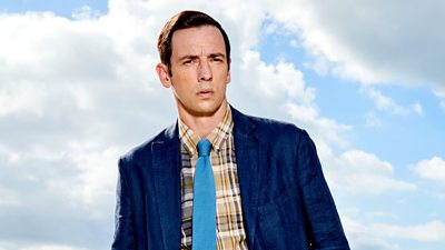 Di Neville Parker (Ralf Little) stood in front of a blue sky. 