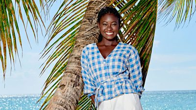 Naomi Thomas (Shantol Jackson) smiling to camera, leaning against a palm tree on a beach. 