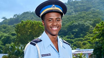 Marlon Pryce (Tahj Miles) stood in uniform. He smiles to camera, a beach and buildings are visible in the background. 