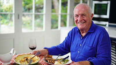 Photo of Rick Stein at a dining table with plates of food in front of him. 