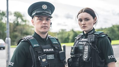 Tommy Foster (Nathan Braniff) and Annie Conlon (Katherine Devlin) are pictured in uniform, wearing protective police vests outdoors