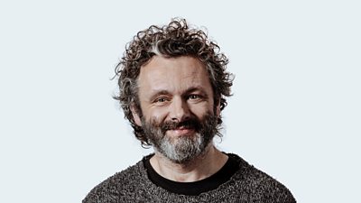 Michael Sheen is pictured smiling. His head and shoulders are visible against a white background. He wears a dark jumper.
