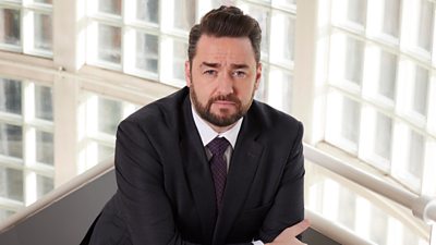 Jason Manford as Mr Savage in Waterloo Road. He stands on a staircase, resting his folded arms over the side. He wears a dark suit and looks serious.