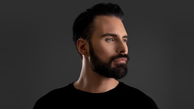 Rylan wears a black top and is pictured side on against a grey background