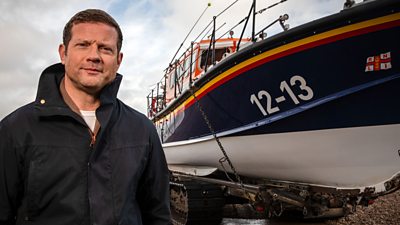 Dermot O'Leary stood in front of a Lifeboat that’s resting on a trailer in a harbour. 