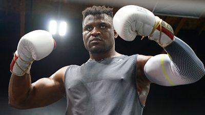 Francis Ngannou in grey training vest holds up both arms in a fighting pose while wearing white boxing gloves