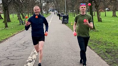 Two men in a park jog towards the camera juggling neon orange and pink balls.