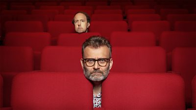 A photo of rows of red theatre seats. Steve Punt and Hugh Dennis rest their heads on the top of the seats 