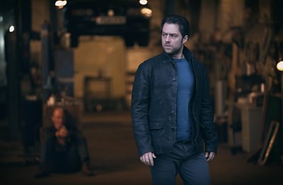 Richard Rankin as John Rebus. He stands in front of a car garage. A blurred figure is visible sitting on the floor behind him.