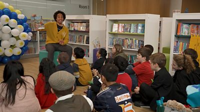 Joseph-Coelho-in-yellow-jumper-reading-to-a-class-of-children-in-a-library