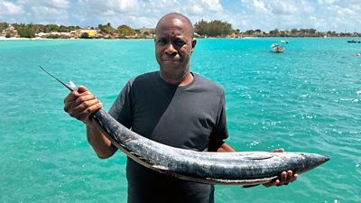 Clive Myrie stands with his back to the sea holding a large fish 