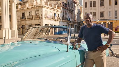 Clive Myrie smiles to camera and leans on a blue classic car 