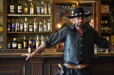 Giovanni se apoya en la barra de un salón y viste ropa de vaquero negro.