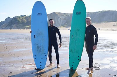 Anton y Gio con tablas de surf en la playa y sonriendo a la cámara