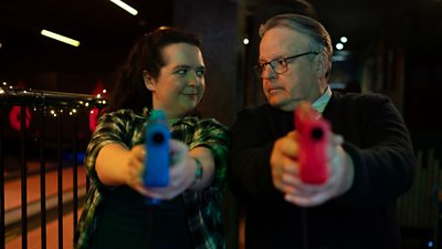 Woman and man looking at each other and pointing red and green carnival toy guns to the camera