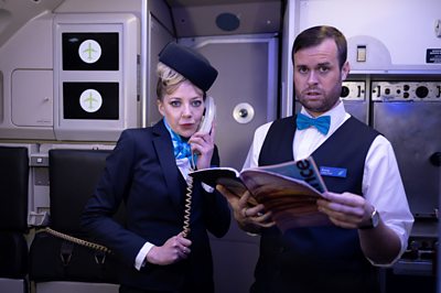 Characters Mandy and Kenny dressed as air stewards. Mandy has a phone to her ear and Kenny is holding a magazine. they're on a plane
