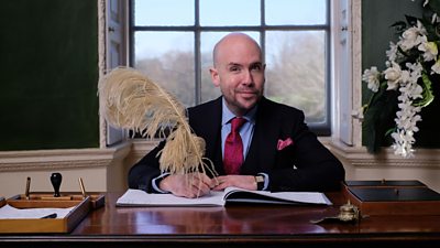 Tom Allen smiles to camera while sat at a desk signing a document with a feathered quill 