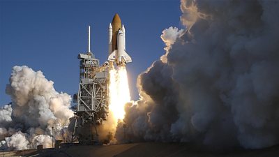 Against a bright blue sky a space shuttle launches in a cloud of smoke