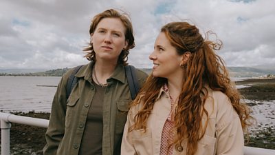 Jade looks into the distance and Collette looks up to Jade smiling. Beach in the background and cloudy sky