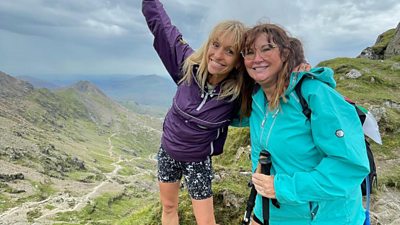 Michaela Strachan and Amanda Lovett smile to camera from the edge of a rocky mountain path. 
