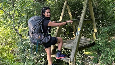 Sonali Shah smiles to camera while climbing over a stile between two fields, surrounded by vibrant, green foliage. 