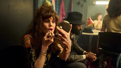 Michelle Dockery as Estella applying lipstick while peering into a small make-up mirror 