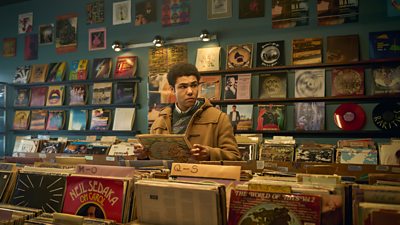 Levi Brown as Dante holding a vinyl cover in a record store. 