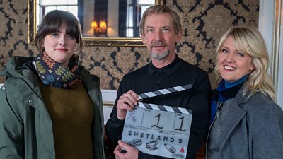 DI ‘Tosh’ Mcintosh (Alison O’Donnell), Euan Rossi (Ian Hart) and DI Ruth Calder (Ashley Jensen) smile to camera. Euan holds a clapperboard that reads: "Shetland 9"