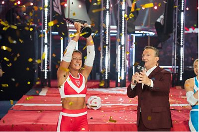 Bradley Walsh speaks into a microphone as the red Gladiators Champion holds a trophy aloft