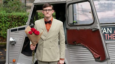 Lorn Macdonald as character Lee. He is standing in front of a van with the door open holding roses with a suit jacket and bow tie 