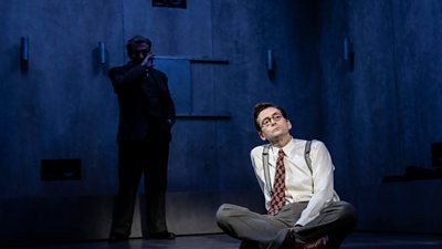 Still from the play good. David Tennant sitting on the floor wearing a shirt and tie looking up pensively. Elliot Levy standing in the background in the shadows