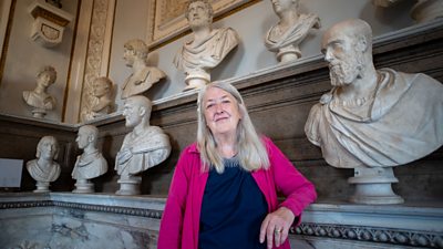 Mary Beard leans against a marble shelf, with busts of multiple figures behind her