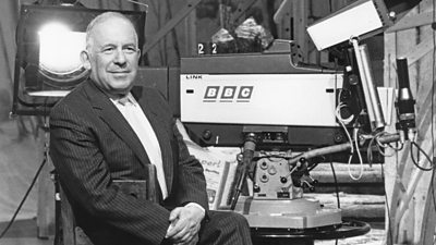 A black and white image of Sir Paul Fox, sitting next to a BBC television camera and studio lights