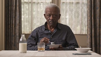 Lennie James as Barrington Walker, sat at a dining table holding a bowl of cereal. A glass tumbler of amber-coloured liquid sits on the table in front of him, alongside a bottle of milk.
