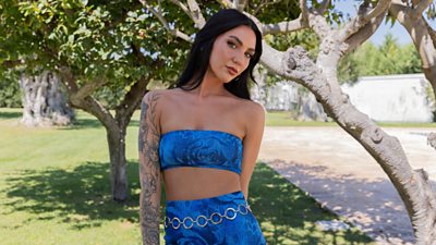 contestant cara looking to camera. she wears a blue tie dye two piece crop top with dark hair and a tattoo sleeve on one arm