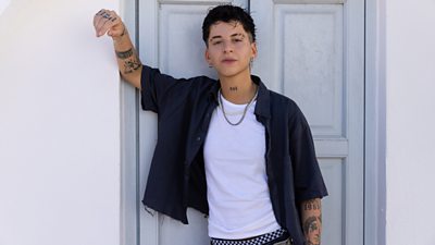 contestant Fiorenza leaning on a doorway. she wears an open short sleeved navy shirt with white tshirt and silver chain. She has short dark hair and tattoos on arms and one on neck