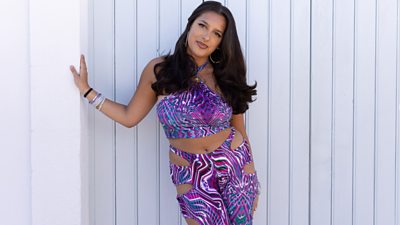 contestant priya leaning on white frame. she wears a purple psychedelic pattern two piece with bits cut out at the top of the trousers. she has long brown hair