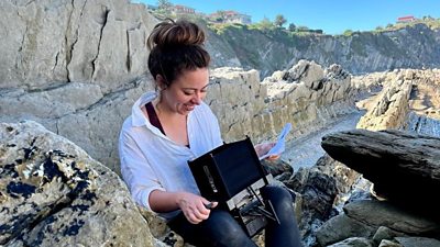 A white woman with brown hair scooped into a bun smiles and looks down, she is wearing a white shirt with rolled up sleeves and is perched on a rocky outcropping, a camera hood in her lap, watching the recording. In the background, a wilderness of grey rocks and green plants.