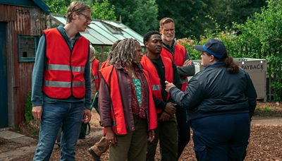 Three men and woman wearing red hi vis jackets while woman wearing navy uniform with cap looks talks to them 