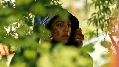Rhianne Barreto as rani with her hood up and cuts to her face looking shocked at something offscreen with leaves in the foregrount