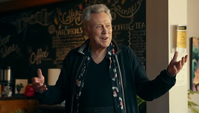 christopher walken as character frank his arms are outspread and he's smiling widely at something offscreen. background is a cafe coffee shop