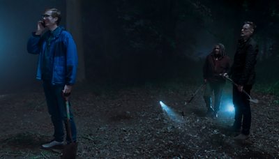 stephen merchant as greg on the phone looking to the left. to the right of him are two people holding shovels with torches looking at him. they look like they're all in a dark forest