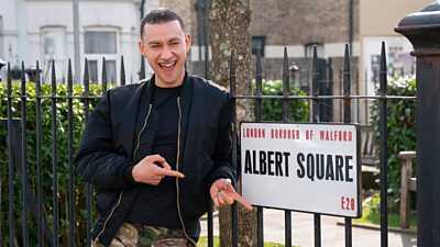Olly Alexander smiles from the set of EastEnders and points to a road sign for Albert Square