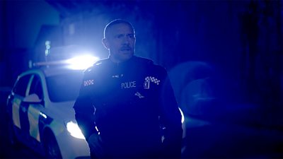 Chris (Martin Freeman) wearing police officer’s uniform, stood in front of a police car. The flashing blue lights of the car illuminate Chris as he looks at something ahead of him. 