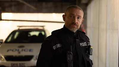 Chris (Martin Freeman) wearing police officer’s uniform, stood in a car park beside a police car. 