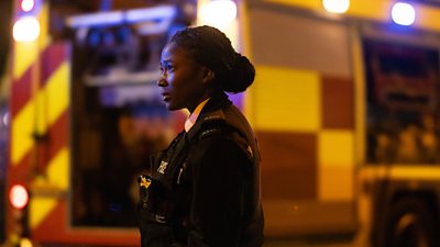 Rachel Hargreaves (Adelayo Adedayo) wearing police officer’s uniform, stood in front of an emergency vehicle with flashing lights. 