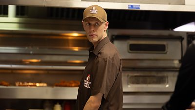 Marco (Josh Finan) stood in the kitchen of a fast-food restaurant wearing a uniform and cap branded with “Krunchy Fried Chicken.”