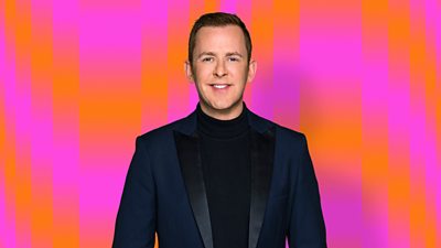 Scott Mills wearing a black top and blazer with an orange and pink background