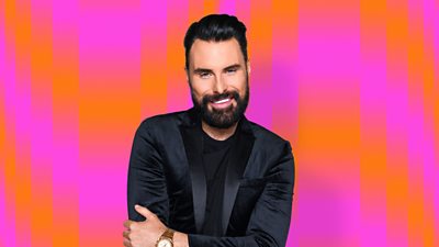 Rylan wearing a black top and blazer with an orange and pink background