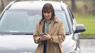 character monty looking at her phone in the rain with car in background